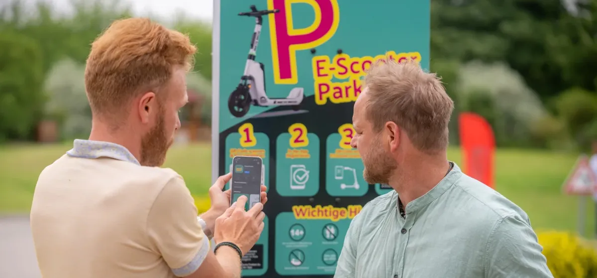 Zwei Männer am Smartphone vor einer E-Scooter-Infotafel